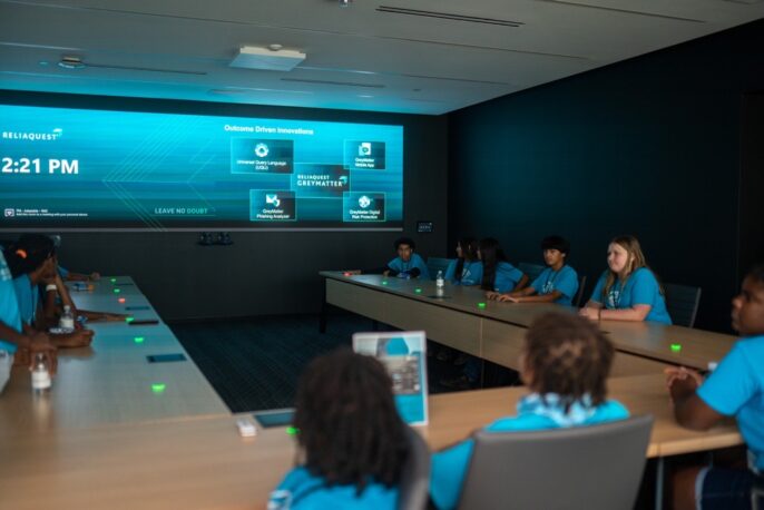 Students sit around a desk discussing cybersecurity innovation at the ReliaQuest offices in Tampa, Florida.